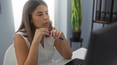 Ofiste çalışan bir kadın, elinde kalem tutarken odaklanmış görünüyor, arka planda bitki ve dosya klasörleri olan ofis malzemeleriyle çevrili..