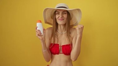 Bikinili kadın, sarı arka planda güneş kremi sürerek başarıyı kutluyor. Gururlu ve heyecanlı. Kollarını kaldırarak zafer kazanıyor.