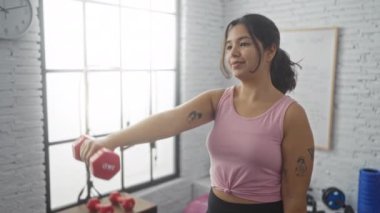 Spor salonunda spor salonunda halterlerle egzersiz yapan İspanyol bir kadın ve arka planda beyaz bir tahta..