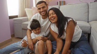 Latin bir aile, kadın erkek ve çocukla oturma odasında oturmuş gülümsüyor ve telefonla konuşuyor.