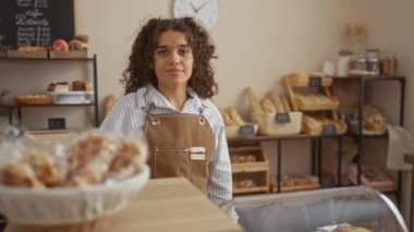 Sıcak bir fırında gülümseyen bir kadın kahverengi bir önlük giyiyor ve arka planda ekmek dolu raflar var..