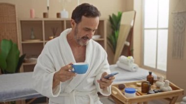 Olgun bir adam bir fincan kahvenin keyfini çıkarırken etrafındaki sağlıklı şeylerle birlikte sakin bir spa odasında akıllı telefonunu kullanıyor.