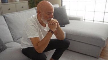 Orta yaşlı, kel kafalı ve sakallı bir adam modern oturma odasındaki bir koltukta düşünceli bir şekilde oturuyor. Düşünce ve derin düşünme duygusu sergiliyor..