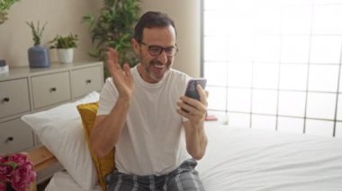 Latin kökenli orta yaşlı bir adam, bitki ve dekorla çevrili, rahat bir yatak odasında yatağında otururken akıllı telefonuyla video konferans yapıyor..