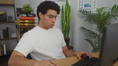 Ofiste çalışan genç bir adam, bilgisayar kullanırken boynu ağrıyan, arka planda kitaplar, bitkiler ve ofis malzemeleri olan bir adam..