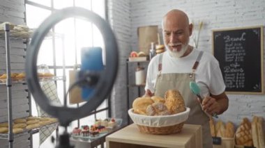 Olgun bir adam kendini bir fırında kaydediyor, elinde bir sepet ekmek tutarken yeni pişmiş mallarla çevrili, kapalı bir mekanda fırın ürünleri sergiliyor.