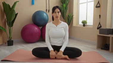 Spor salonunda meditasyon yapan kadın paspasın üzerinde oturuyor. Etrafı spor aletleri ve bitkilerle çevrili.