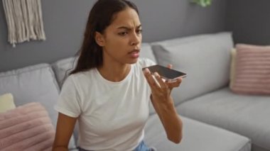 Telefonda öfkeli bir ifadeyle konuşan bir kadın oturma odasındaki kanepede oturan beyaz tişörtlü bir kadın.