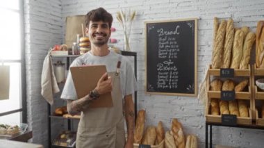Sakallı ve dövmeli genç bir adam elinde bir pano ve fırında duruyor ve duvarda bir menü dolusu ekmek gösteriyor.