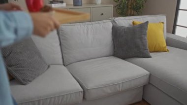 Bıyıklı genç adam kahve ve kek yiyor modern oturma odasında, gri kanepeli ve sarı yastıklı.