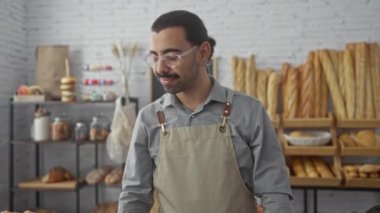 Fırında çalışan bıyıklı genç bir adam gözlüklü ve önlüklü, modern bir iç mekanda taze ekmek raflarıyla çevrili..