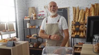 Gri saçlı ve sakallı yakışıklı bir adam ekmek ve hamur işleriyle dolu bir pastanede kolları çapraz pozlar veriyor. Önlük takıyor ve güvenli bir şekilde rafların önünde duruyor..