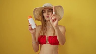 Bikinili beyaz kadın elinde güneş kremi tutuyordu. Seni ciddi bir hareketle gösteriyordu. Sarı arka planda izole edilmişti.