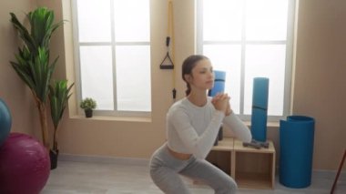 Spor salonunda spor aletleriyle egzersiz yapan bir kadın. Arka planda spor topları ve bitkiler de var.