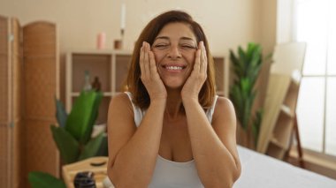 İspanyol kadın spa 'da rahatlatıcı bir anın tadını çıkarıyor, elleri yüzünde, bir sağlık merkezinin kapalı ortamında gülümsüyor..