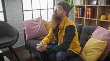 Düşünceli, sakallı, gözlüklü ve sarı ceketli bir adam gri bir kanepede oturuyor, renkli yastıklarla çevrili..