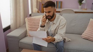 Oturma odasındaki koltukta oturan sakallı genç adam, telefonda konuşurken endişeli görünüyor ve elinde kağıtlar var..
