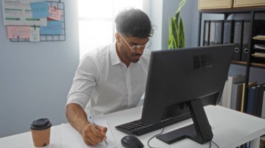 Geniş bir masaüstü bilgisayarıyla özenle çalışan genç bir İspanyol, organize dosyalar, bir fincan kahve ve duvardaki motivasyon notlarıyla çevrili.