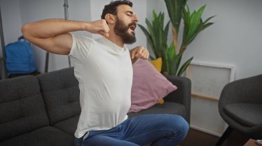 Tired bearded man in casual attire yawning and stretching on the couch in a modern living room setting. clipart