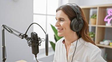 Kulaklıklı orta yaşlı bir kadın, podcast stüdyo ortamında mikrofona konuşuyor, profesyonellik ve pozitiflik yayıyor..