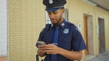 Afro-Amerikan polis memuru şehir sokaklarında akıllı telefon kullanıyor.