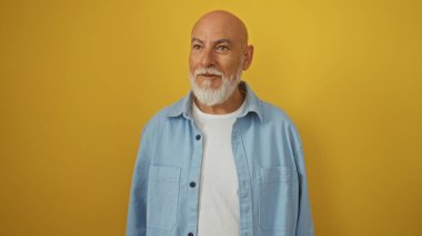 Gri saçlı ve sakallı yakışıklı olgun bir adam izole edilmiş sarı bir arka plana gülümsüyor.