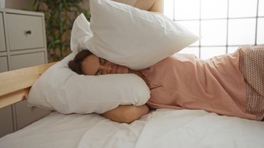 Parlak bir yatak odasında, yastıkla kulaklarını kapatan bir kadın gürültüden rahatsız görünüyor..