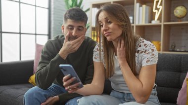 Endişeli bir çift, modern bir oturma odasında, ilişki dinamiklerini yansıtan bir akıllı telefonu birlikte inceliyor..