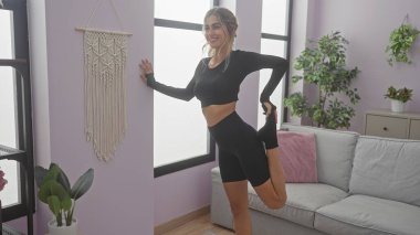 Aktif kıyafetli gülümseyen genç bir kadın, bitki ve duvar dekoruyla modern bir oturma odasında esneme egzersizi yapıyor..