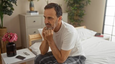 İspanyol bir adam yatak odasında bir yatakta oturuyor. Dekoratif bitkiler ve mobilyalarla dolu bir ev ortamında düşünceli ve düşünceli görünüyor..