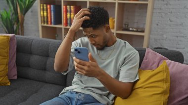 Düşünceli Afrikalı adam, rahat bir oturma odasında gri koltukta renkli yastıklarla telefona bakıyor.