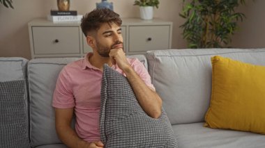 Sakallı genç İspanyol bir adam, rahat bir oturma odasında kanepede, elinde yastıkla, ev dekoruyla düşünceli bir şekilde oturuyor..
