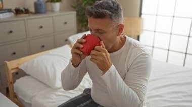 Orta yaşlı bir adam rahat mobilyaları ve dekorasyonu olan rahat bir yatak odasında kırmızı bardaktan su içiyor.