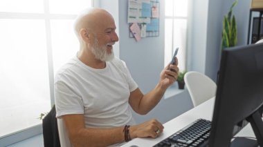 Ofiste oturmuş akıllı telefonuna bakarken gri sakallı, gülümseyen orta yaşlı bir adam.