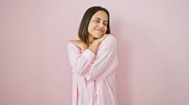 Pembe gömlekli genç İspanyol kadın kapalı gözlerle poz veriyor ve solgun pembe bir arka plana gülümsüyor..