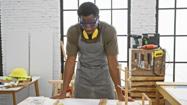 Bir atölyede odaklanmış bir adam, marangozluk istasyonundaki aletlerle çevrili ahşap malzemeleri inceliyor..