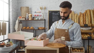 Genç, sakallı, kahverengi önlük ve plastik eldiven giyen, fırının içinde pembe bir pasta kutusu hazırlayan İspanyol bir adam.