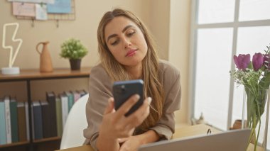 Genç bir kadın, modern dekor ve doğal ışıkla çevrili rahat bir ev ortamında akıllı telefon kullanıyor..