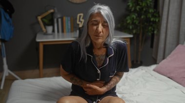 Yaşlı bir kadın, gri saçlı, yatak odasında bir yatakta oturuyor, pijama giyiyor ve acı çekiyor gibi görünüyor, arka planda modern bir ev var..