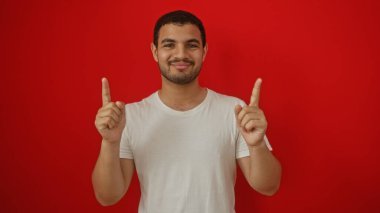 İzole edilmiş kırmızı arka planda poz veren genç İspanyol adam işaret parmaklarıyla işaret ediyor, beyaz tişört giyiyor, güvenle gülümsüyor.