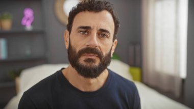 Düşünceli, sakallı, orta yaşlı bir adam iyi döşenmiş bir yatak odasında oturur, incelikle düşünceli bir şekilde ifade eder.