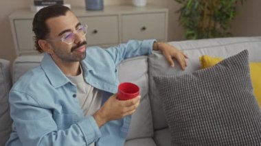 Bıyıklı genç bir adam oturma odasındaki koltukta elinde kırmızı bir bardakla oturur, gözlük takar ve arka planda ev dekoru olan günlük kıyafetler giyer.