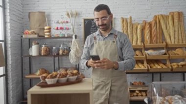 Adam akıllı telefonuyla kapalı bir fırında kek fotoğrafı çekiyor, arka planda ekmek ve hamur işleri rafları var..