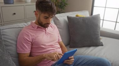Genç adam evdeki rahat atmosferi gösteren yastıklarla çevrili, modern bir oturma odası ortamında tablet kullanarak evde uzanıyor..