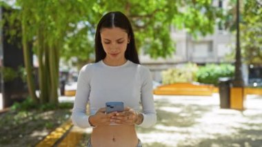 Güneşli bir günde, ağaçlar ve yeşilliklerle çevrili şehir parkında akıllı telefon kullanan genç bir kadın açık hava ortamının keyfini çıkarıyor.