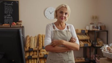 Fırında çalışan bir kadın, etrafı ekmek ve pastalarla çevrili bir şekilde arka planda saatle kapalı dükkan ayarlarını gösteriyor.