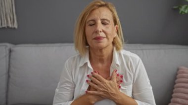 Kadın, oturma odasında elleri göğsünde, huzurlu bir iç mekandaki farkındalığa odaklanmış bir şekilde kendini huzurlu hissediyor.