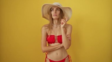 Bikini ve yaz şapkalı beyaz kadın endişeli ve gergin görünüyor, bir soru üzerine derin düşüncelere dalmış, sarı bir arka planda izole edilmiş.