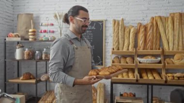 Bıyıklı ve elinde kruvasan tepsisi olan genç adam fırının içinde ekmek ve hamur işleriyle gülümserken.