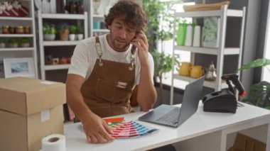 Masadaki renk örneklerini incelerken telefon ve dizüstü bilgisayar kullanan genç bir adam.